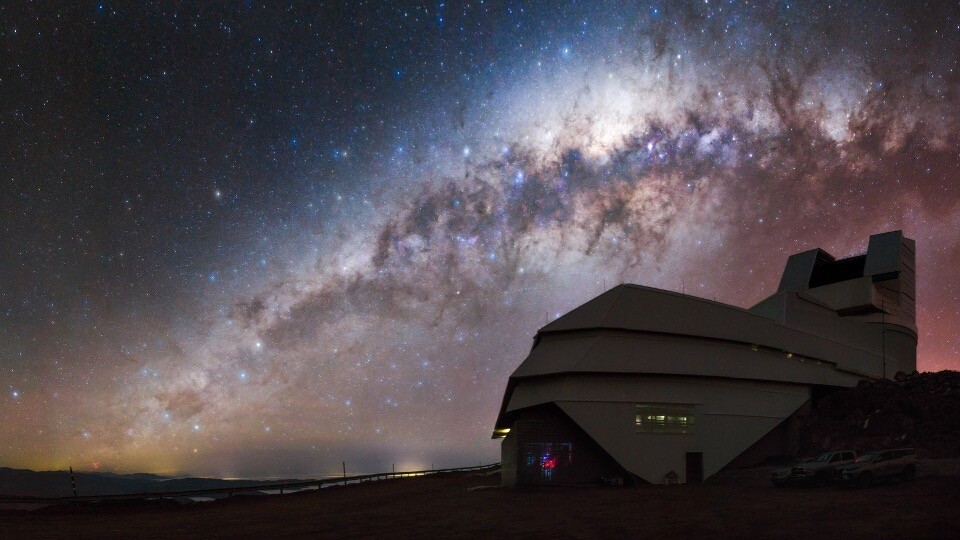 A telescope dome on a mountain ridge beneath the arc of the Milky Way